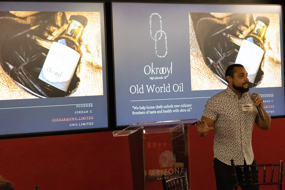 Person presenting at a podium with two projected screens behind them displaying advertisements for "Okroyl Old World Oil." The screens show images of an oil bottle.