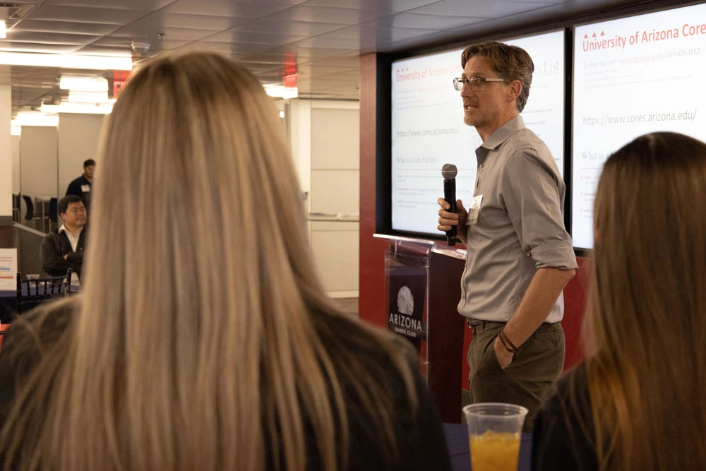 A person presenting at a University of Arizona event, standing with a microphone in front of a projected screen, addressing an audience.