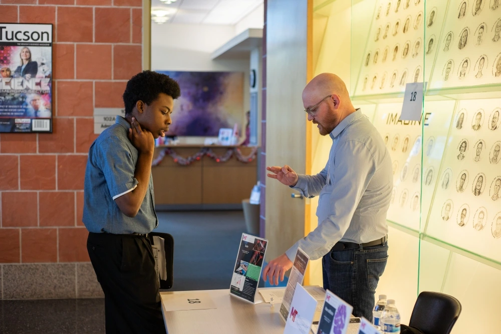 Two individuals discussing over a desk in an office environment, with posters on the wall including one reading "Tucson.
