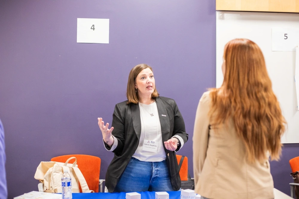Person at a networking event, speaking animatedly to another attendee at a table with papers and cups, in a room with numbered stations on the walls.