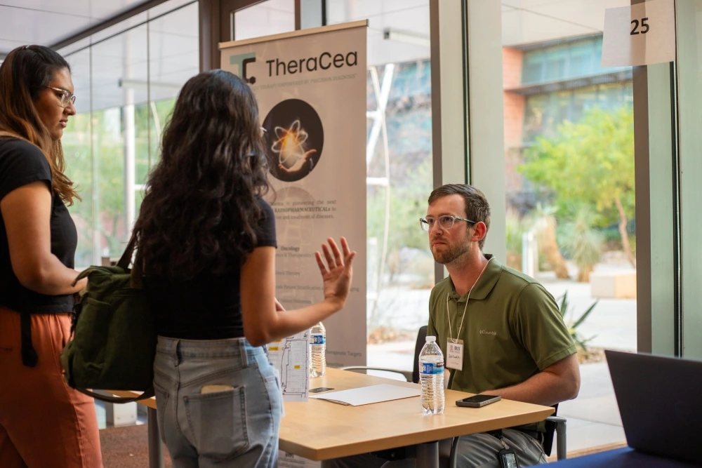 Three individuals discussing at a TheraCea exhibit booth inside a building with large windows and modern architecture.