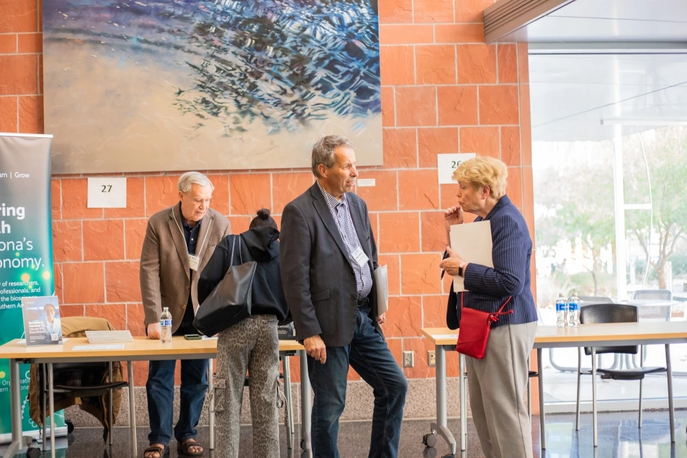 Three individuals are engaged in a conversation at a conference beside numbered booths, under a large abstract painting.