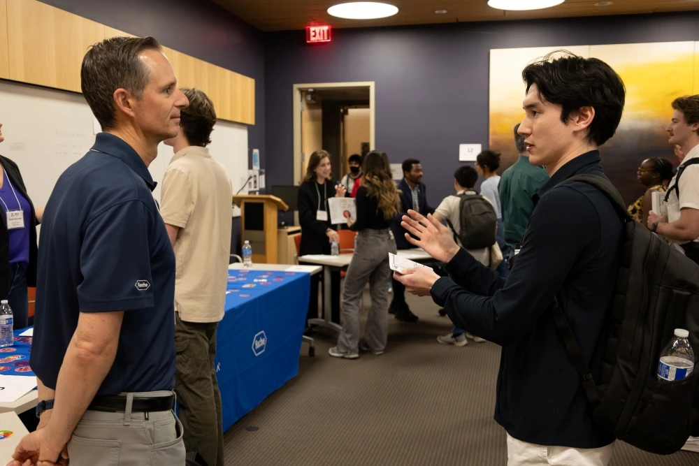 Two individuals are engaged in a conversation at a career fair with a Roche banner visible in the background. Other attendees are interacting and networking around them in a room with vibrant decor.