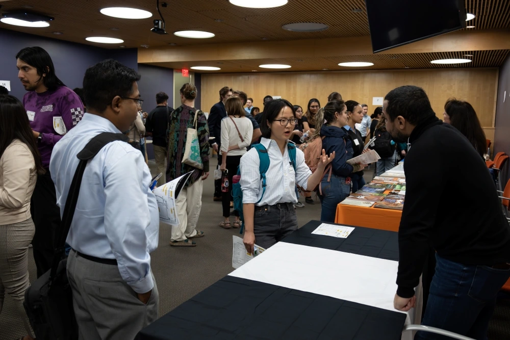 Attendees engaging with a presenter at a conference booth inside a room with wood-paneled walls and informational materials displayed on the table.