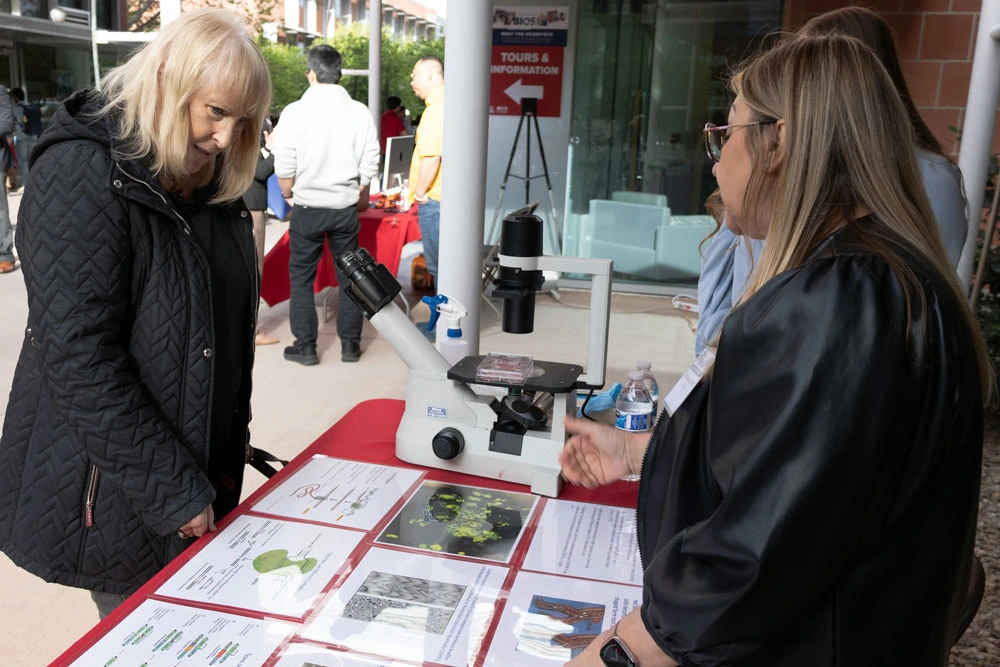 Two individuals are engaged in conversation at an information booth, which features a microscope and various educational materials on display, including maps and images of cells at an outdoor public event.