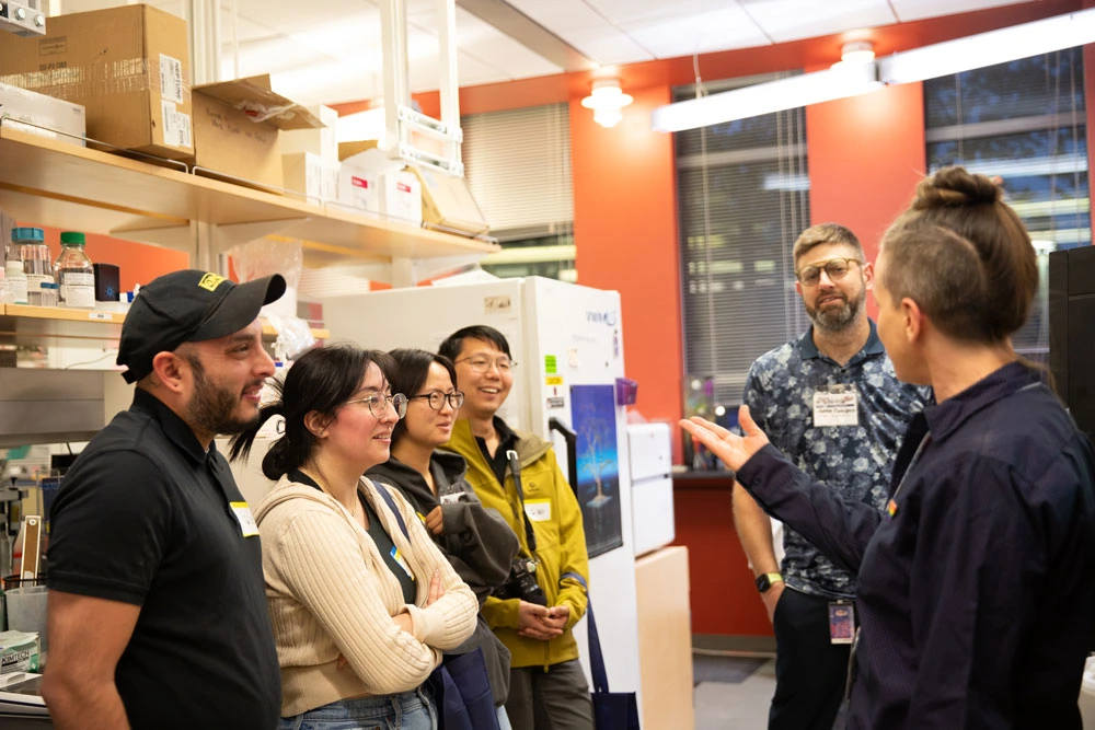 Five individuals engaged in a conversation in a laboratory, with shelves filled with boxes in the background.