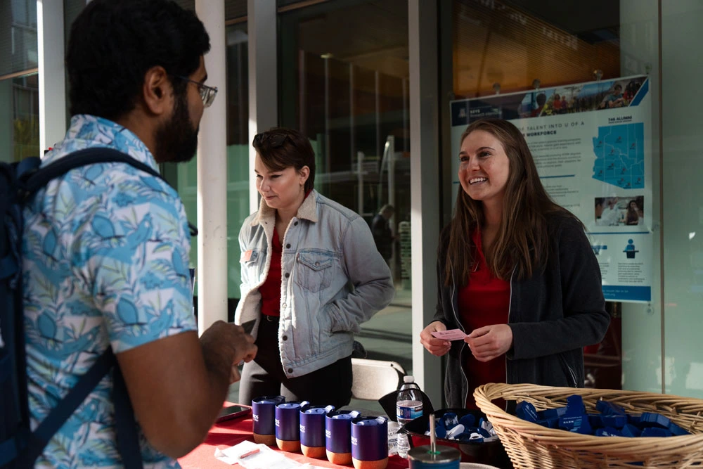 Three individuals engaging in conversation at an informational booth with brochures and promotional items on the table.