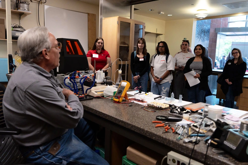 A group of people attentively listens to an instructor at a desk filled with technical equipment and tools.