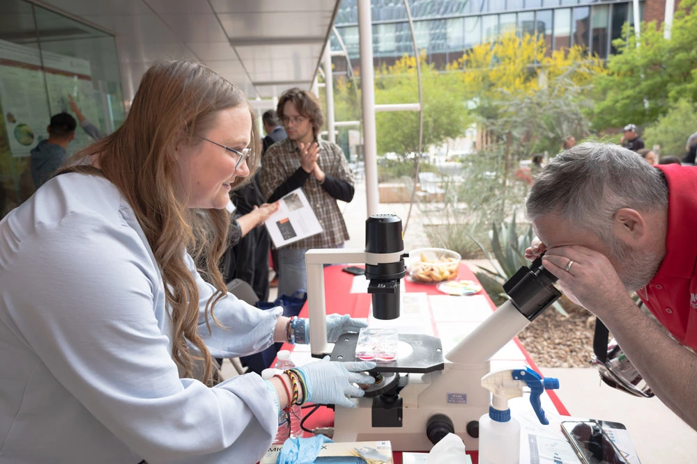 Two individuals using a microscope at an outdoor science exhibit, with one person assisting the other in viewing a sample. There are trees and a building in the background.