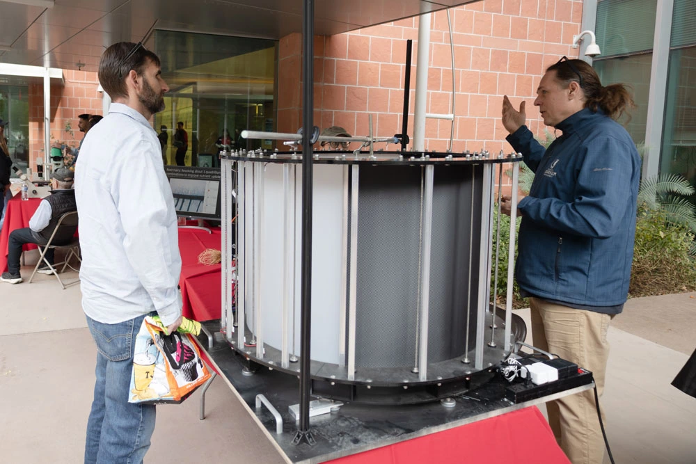 Two individuals conversing beside a large cylindrical scientific apparatus at a public display.