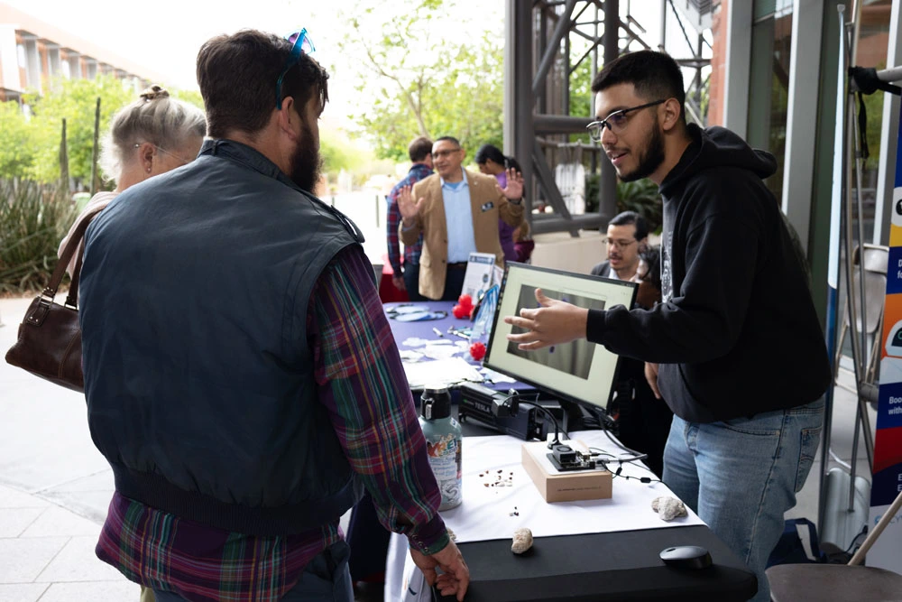 A group of individuals engaged in conversation at an outdoor educational booth with various items displayed on the table, including a monitor and some mechanical parts.