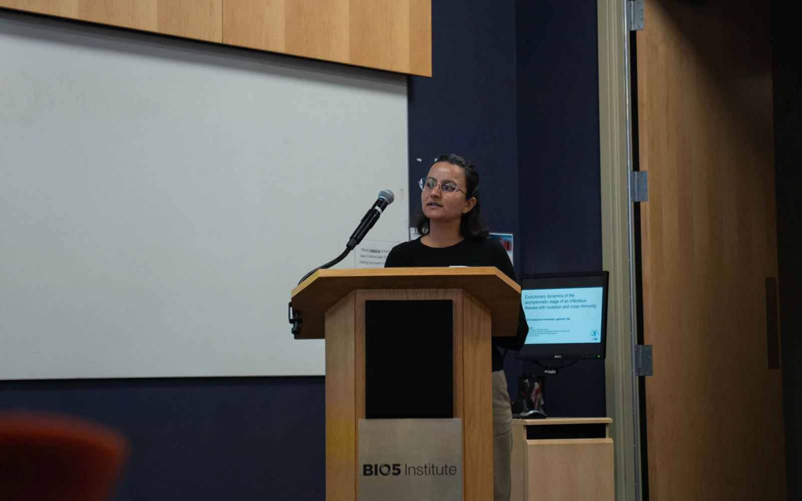 A person stands at a podium labeled "BIO5 Institute" in a lecture hall, facing an audience.