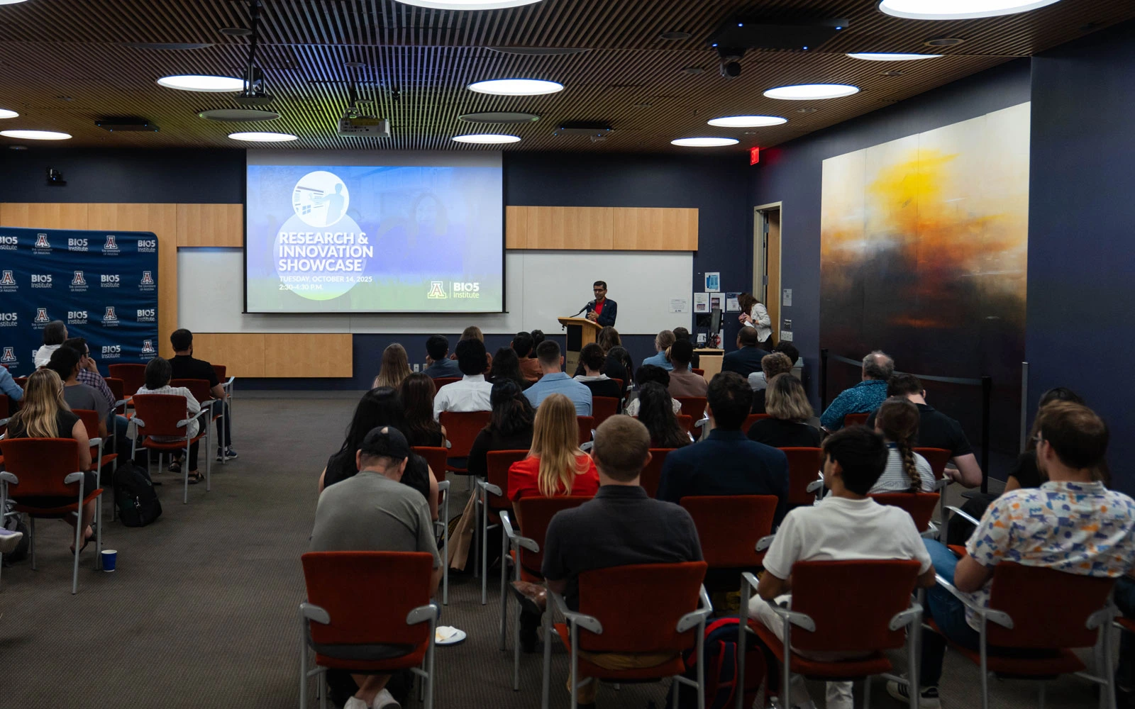 A person presenting at a Research and Innovation Showcase in a university auditorium filled with attentive audience members.