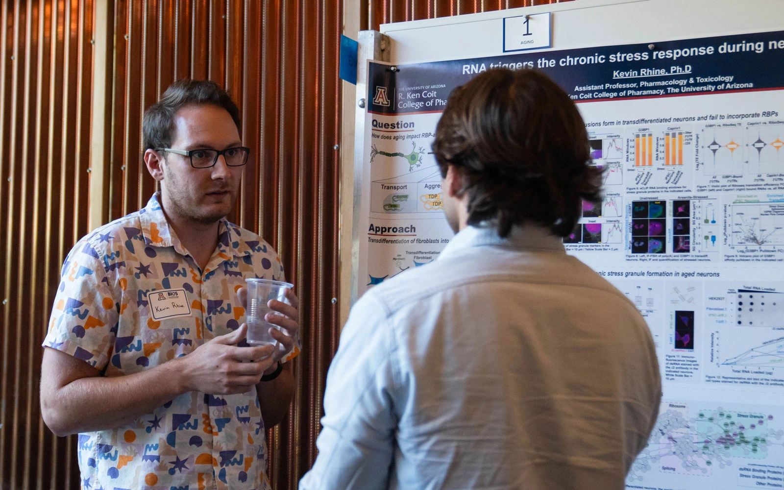 Two individuals engaging in a discussion in front of a scientific poster presentation at a conference. One person is holding a glass of water, and the poster includes various charts and data related to RNA research on stress response.