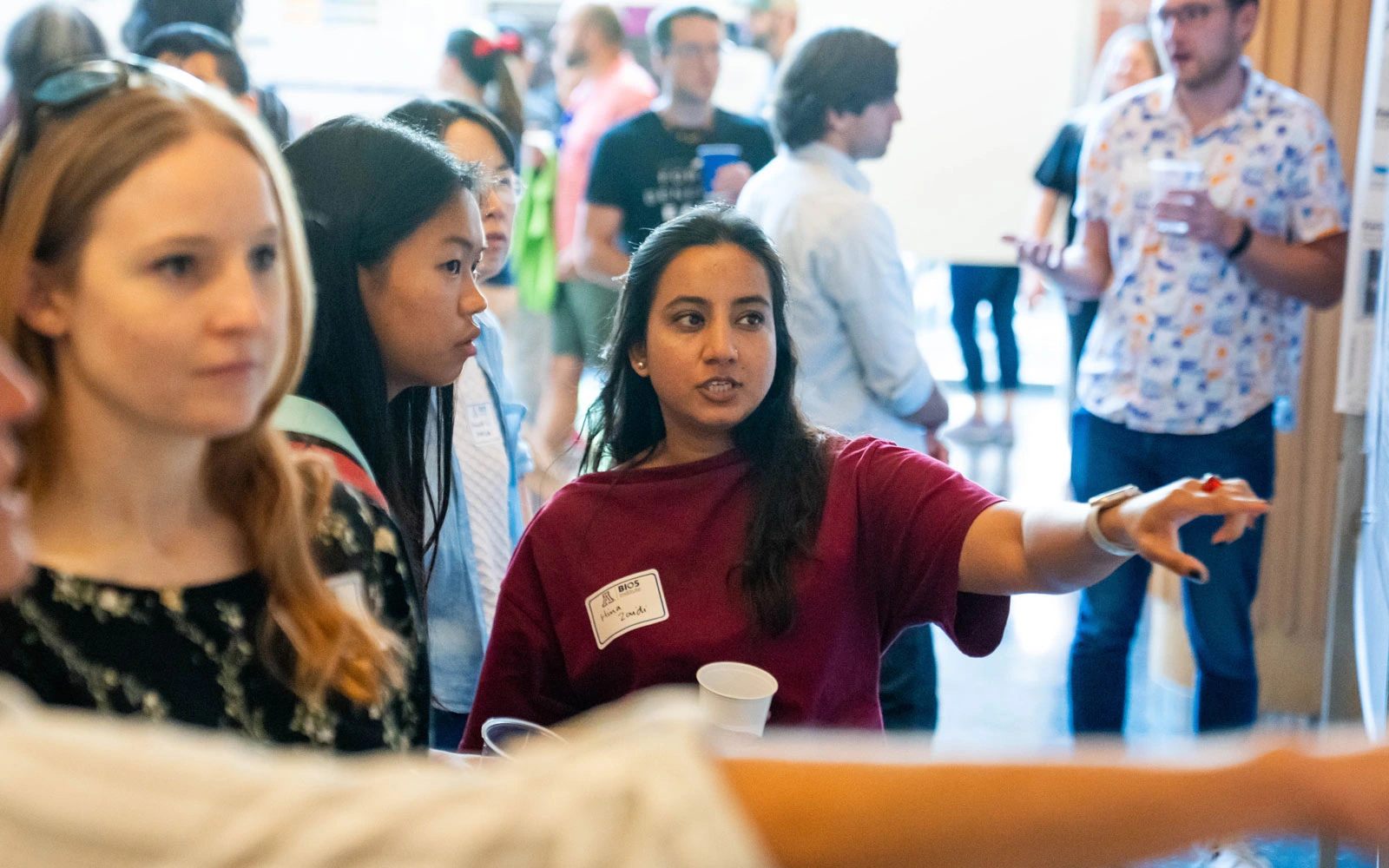 A group of individuals with name badges engaged in a conversation in a crowded event hall. One person is gesturing while speaking to others.
