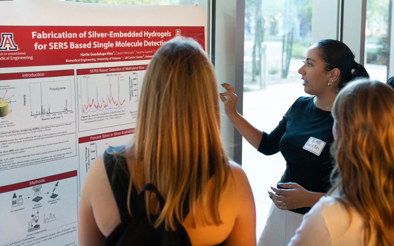 Two individuals examine a research poster titled "Fabrication of Silver-Embedded Hydrogels for SERS Based Single Molecule Detection" at a University of Arizona event. One individual is presenting, pointing at a section of the poster, while the other listens attentively.