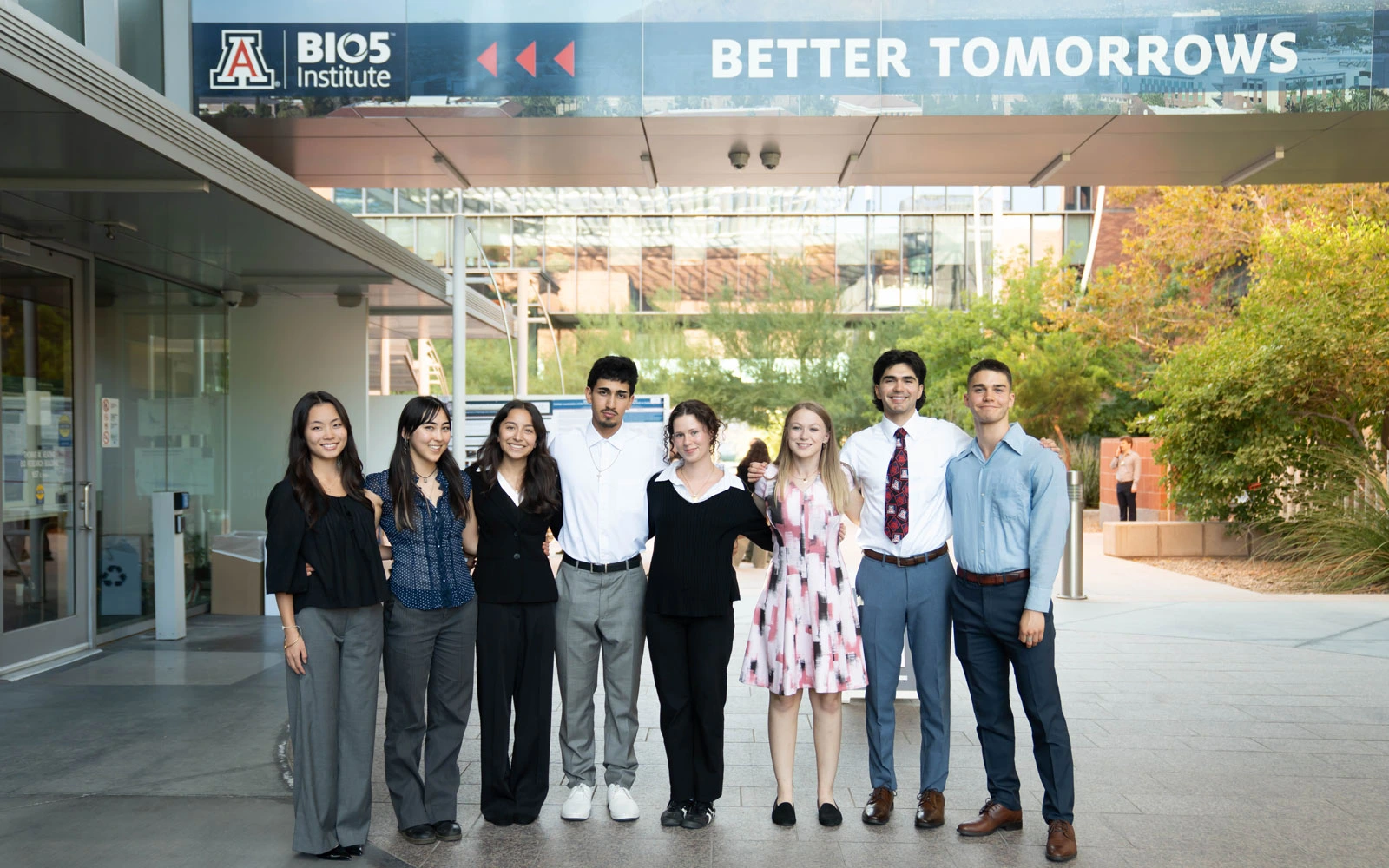 A group of eight diverse individuals smiling and posing in front of the BIO5 Institute, under a sign that says "BETTER TOMORROWS". They are dressed in business casual attire.