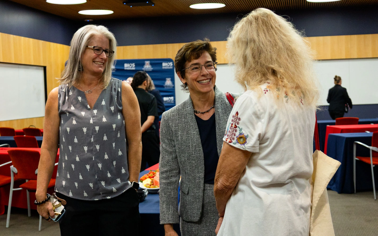Three individuals engaging in a cheerful conversation at a professional event, with snacks visible on a table in the background. 