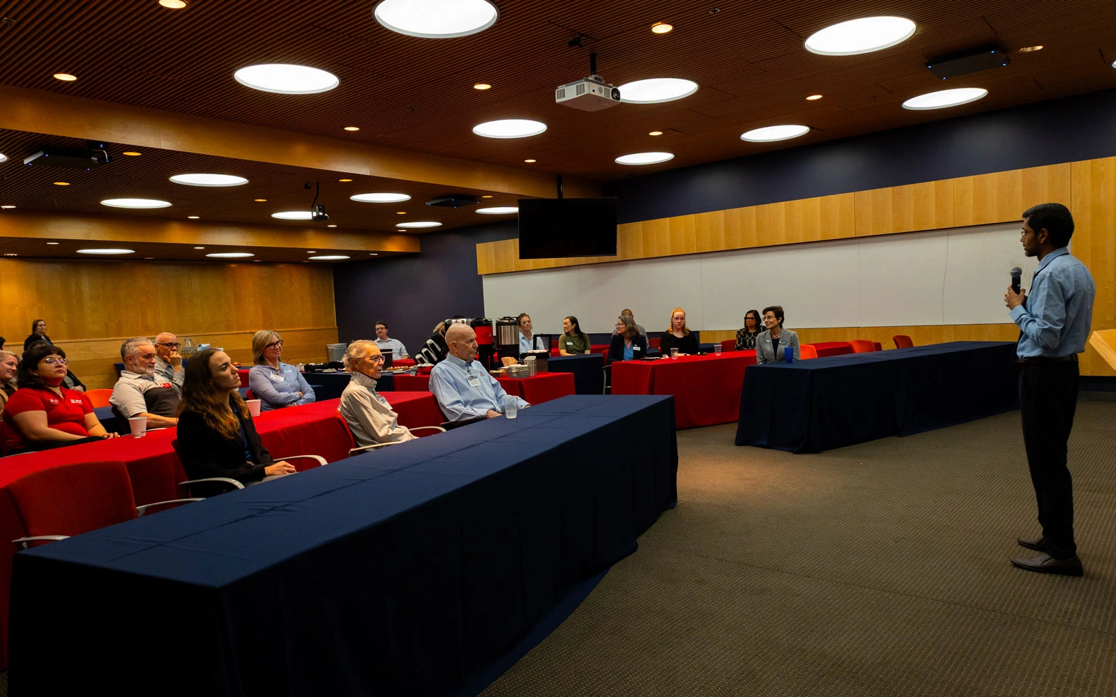 A presenter stands at the front of a lecture hall addressing an audience seated in red chairs. The room is well-lit, with a modern design.