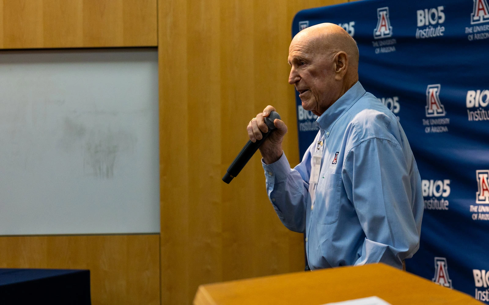 A person is speaking into a microphone at a lectern with the University of Arizona and BIO5 Institute logos displayed in the background.