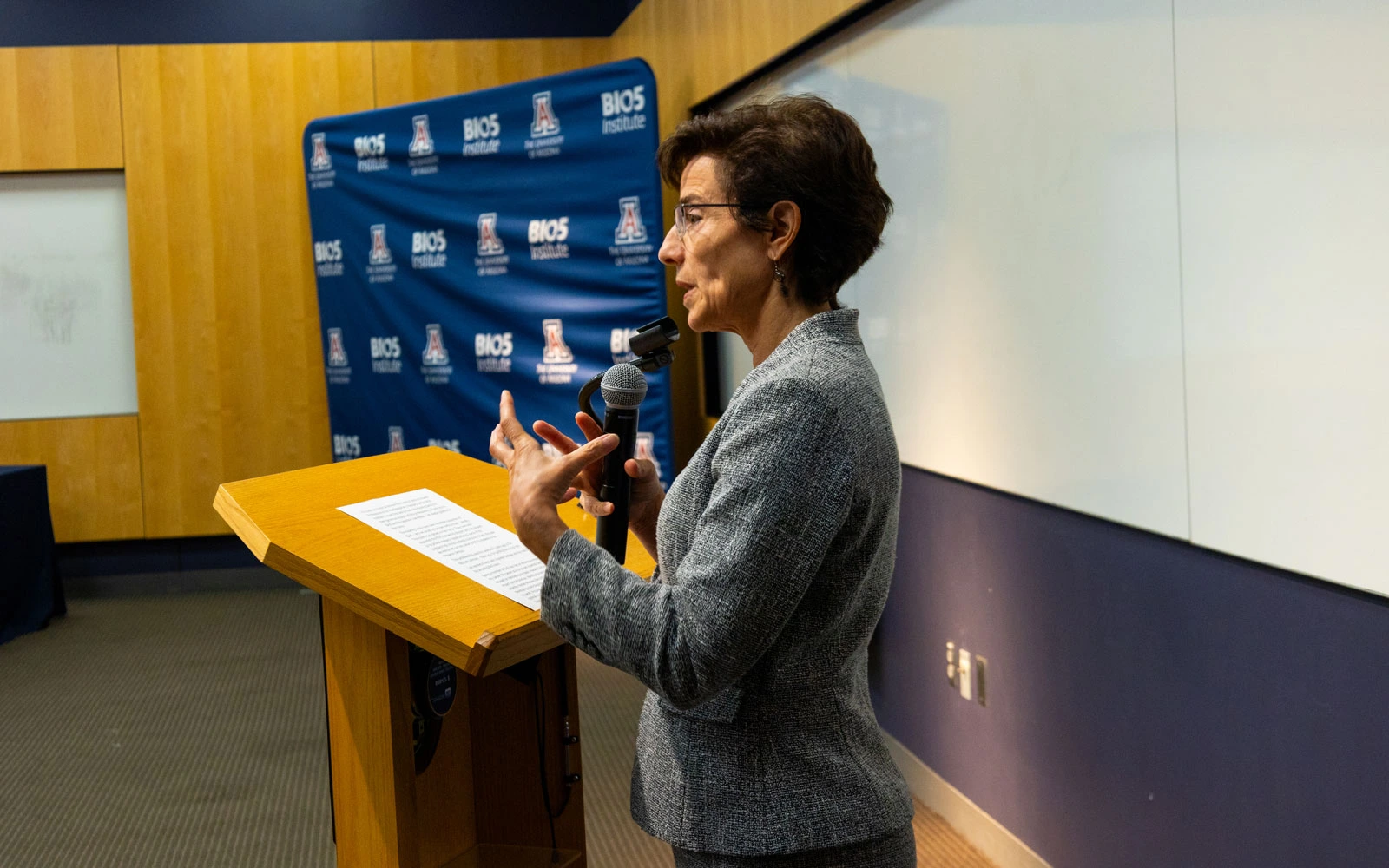 A person giving a speech at a podium with a microphone in a room with a "BIO5" banner in the background.