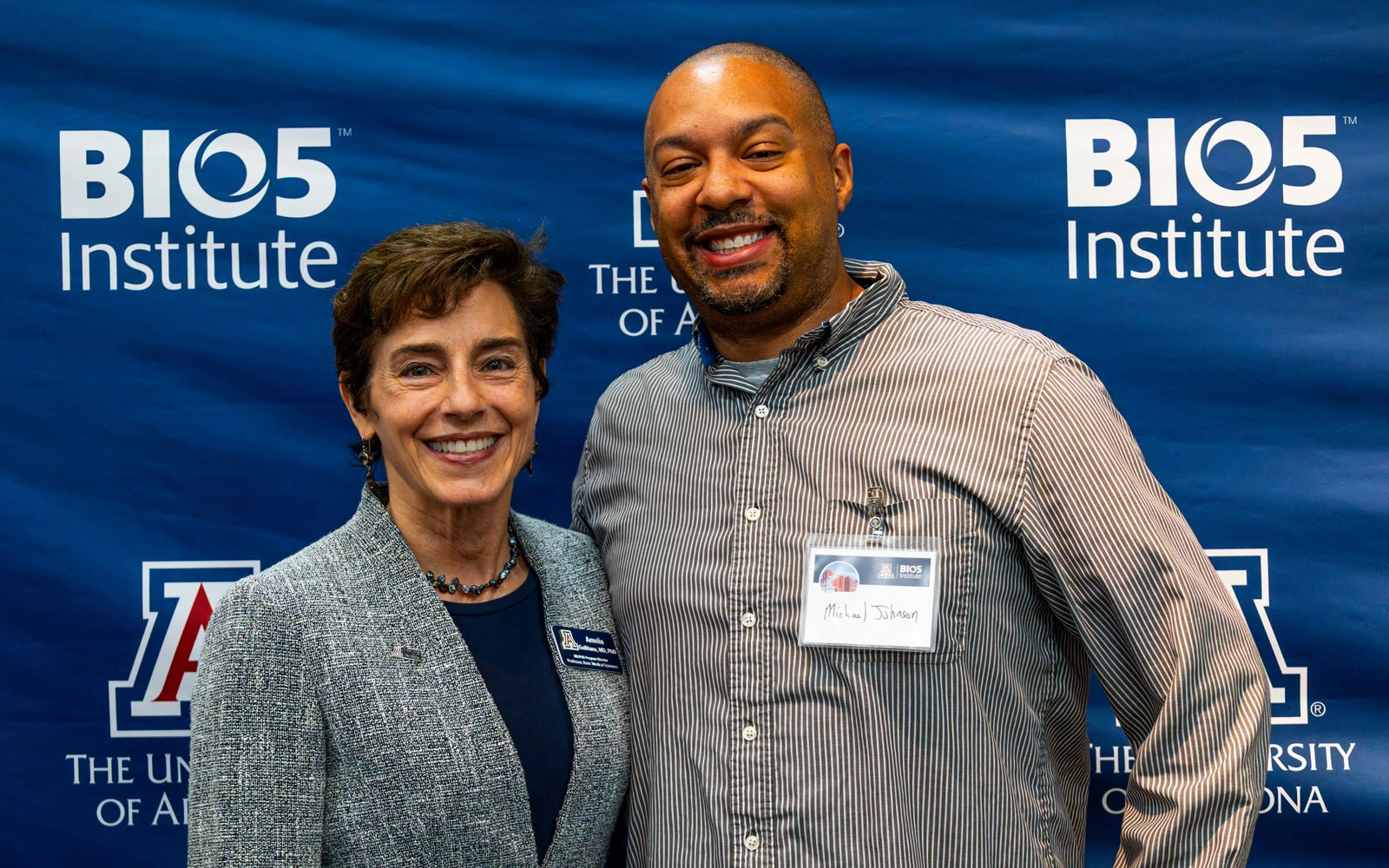 Two individuals smiling at the camera in front of a BIO5 Institute banner at the University of Arizona. One wears a gray blazer, and the other is in a striped shirt with a name tag.