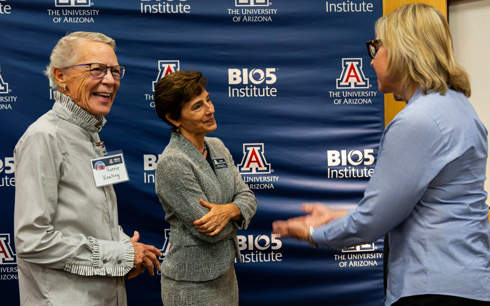 Three individuals are engaging in conversation at the University of Arizona BIO5 Institute, as indicated by the logo-filled backdrop. Two are listening intently while the third, facing away, gestures during the discussion.