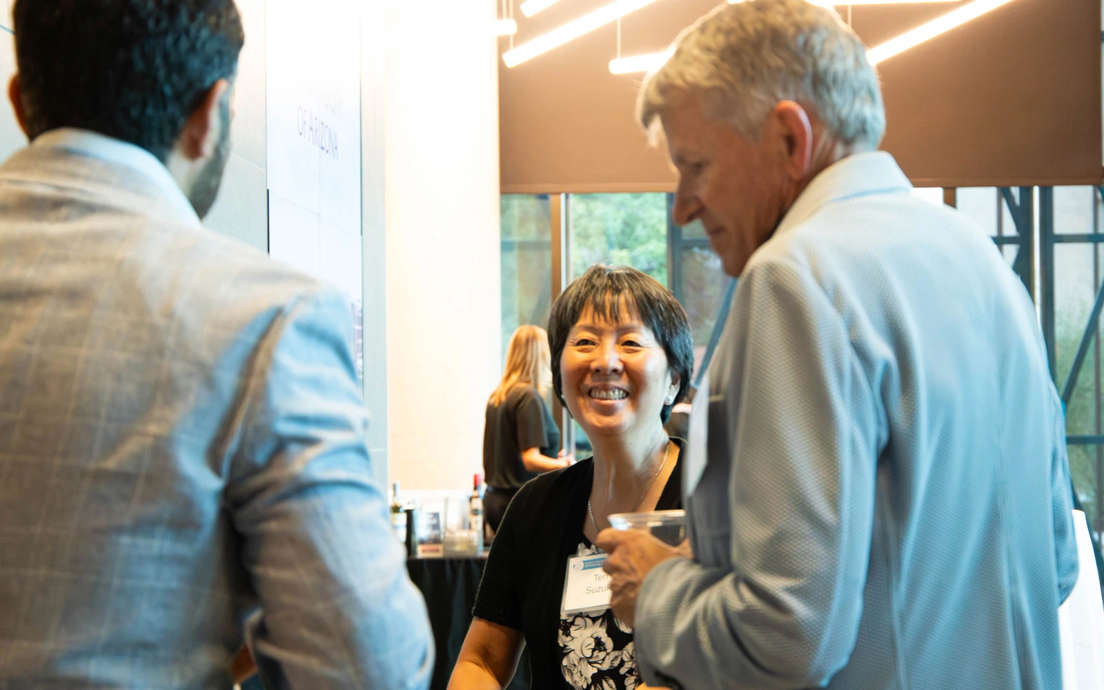 Three professionals conversing happily at a networking event, one of whom is wearing a name tag. The environment features modern architectural elements.