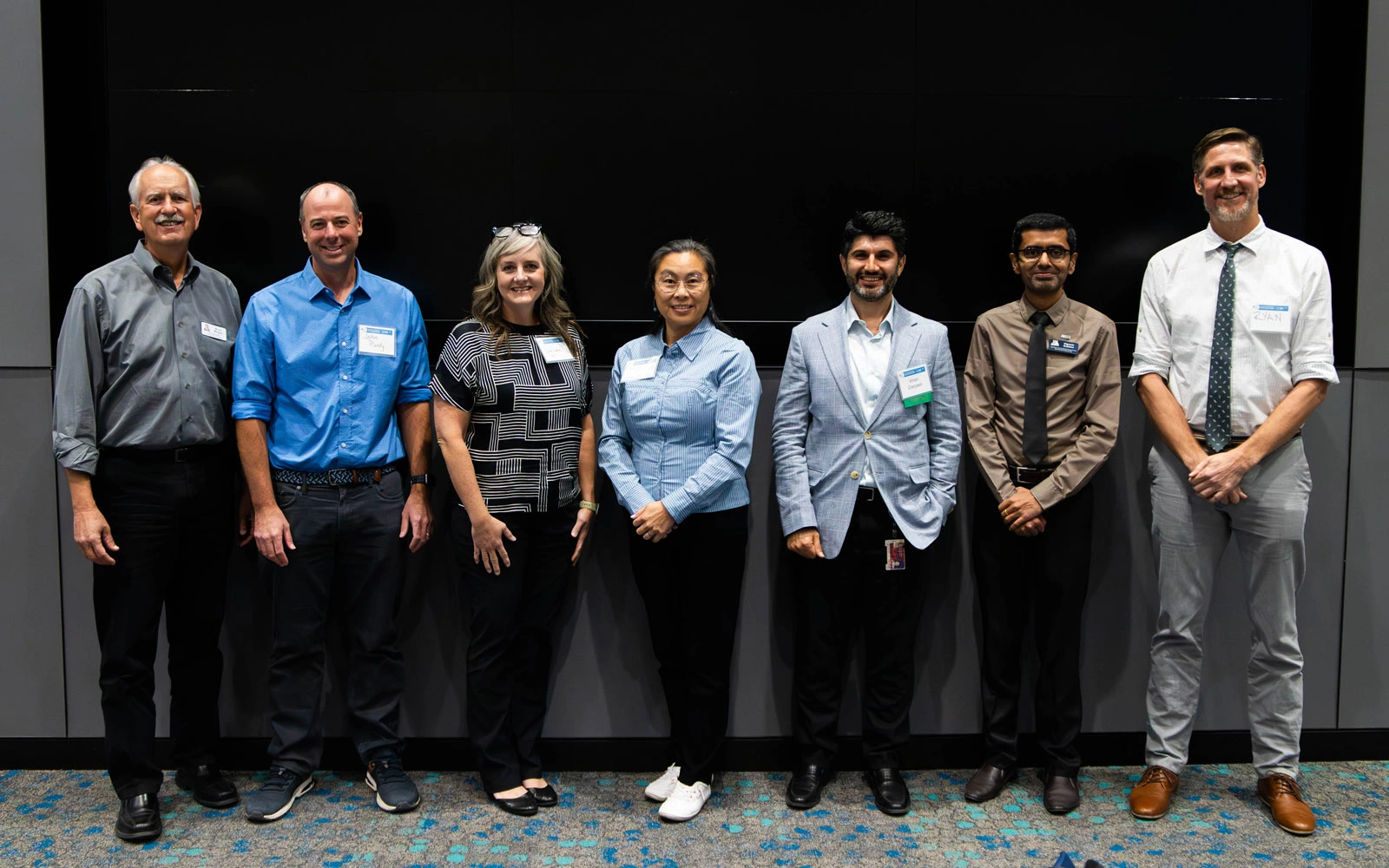 Group of seven professionals with name tags standing in front of a plain black background, smiling.