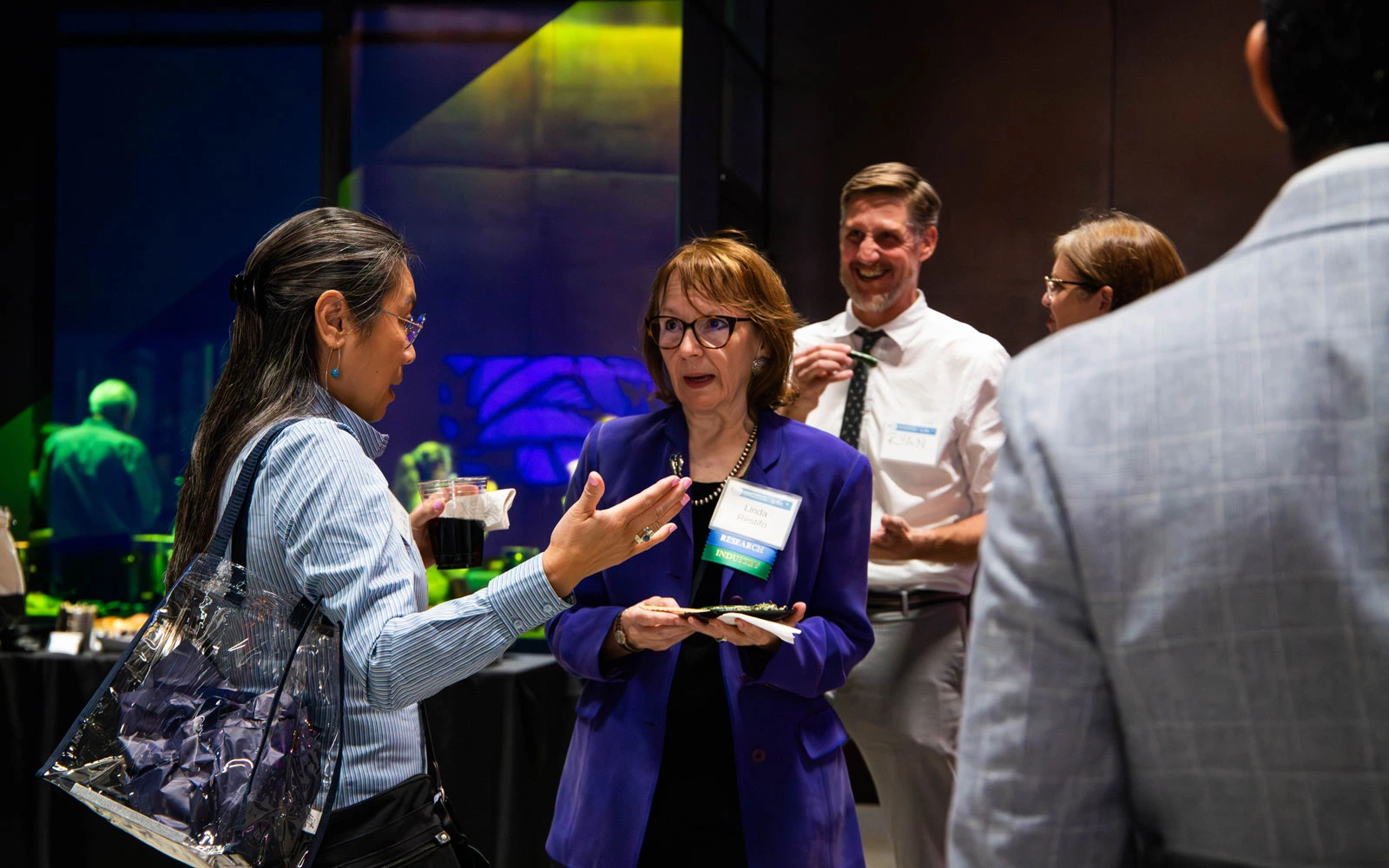 Group of professionals engaging in a conversation at a networking event, with one individual holding a plate of food and a business name tag visible.