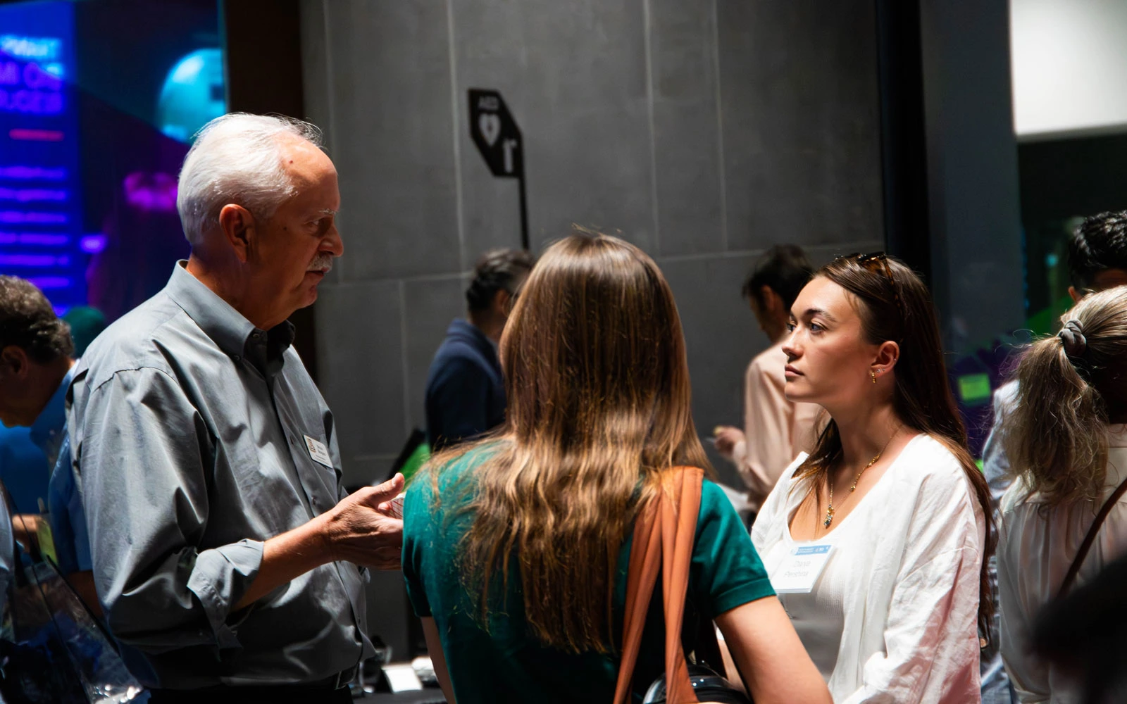 Two people engaging in a conversation at a networking event, with others in the background and ambient lighting.