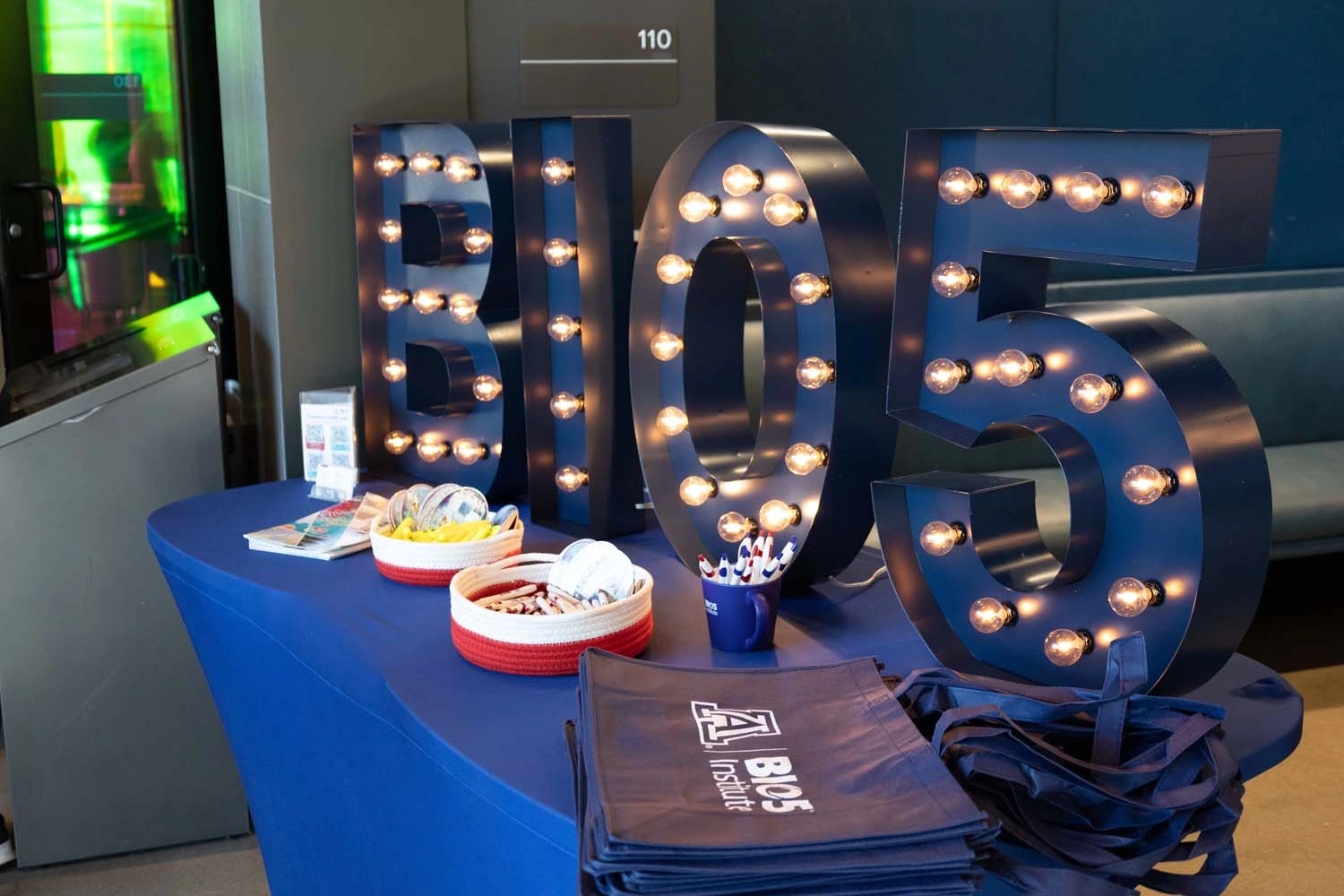 A table displays large illuminated letters spelling "BIO5." On the table are brochures, two baskets with items, a container with pens, and a stack of BIO5 Institute branded bags.