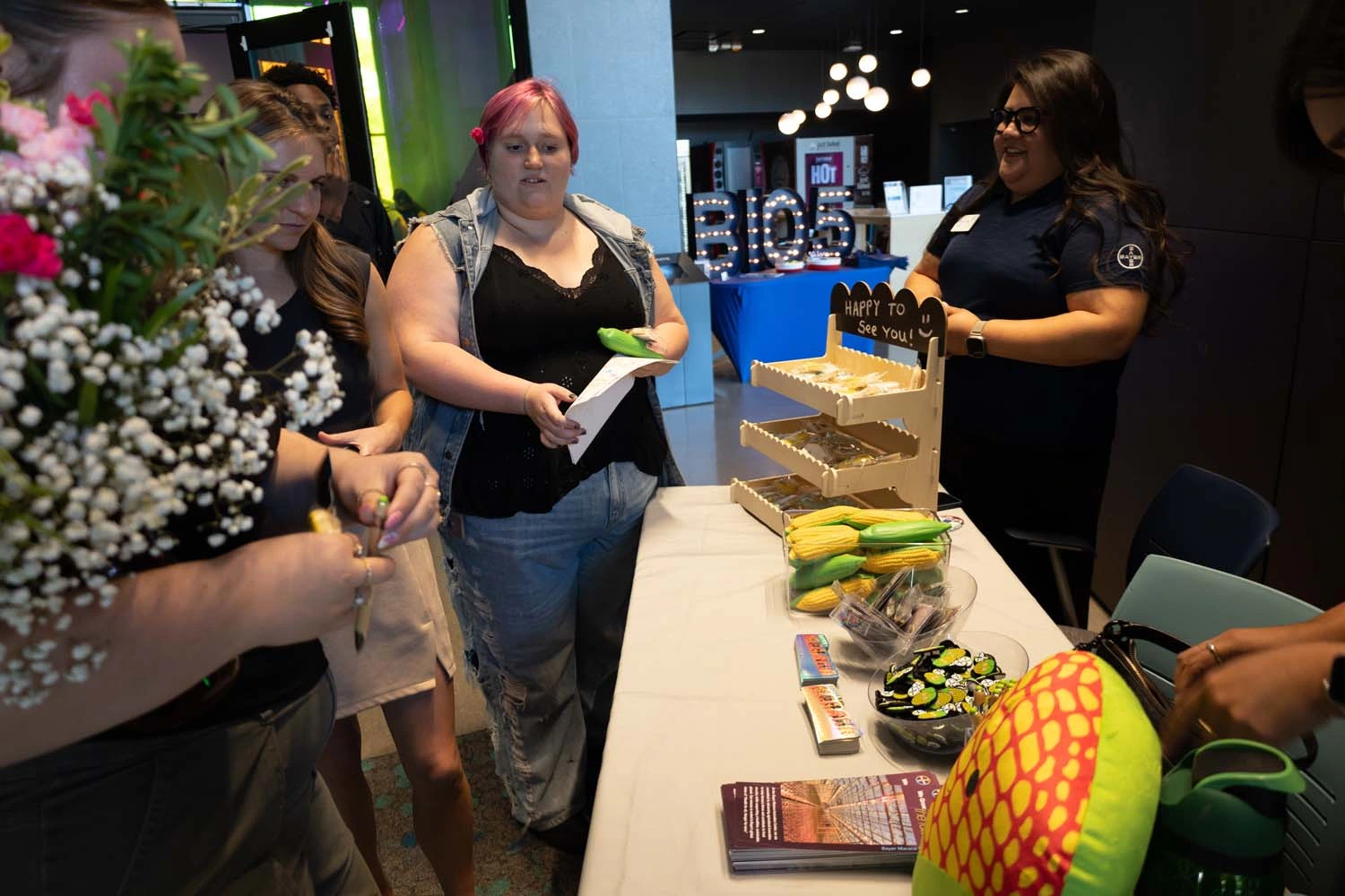 A group of people stands around a table at a convention, where various items like corn-shaped stress toys and brochures are displayed.