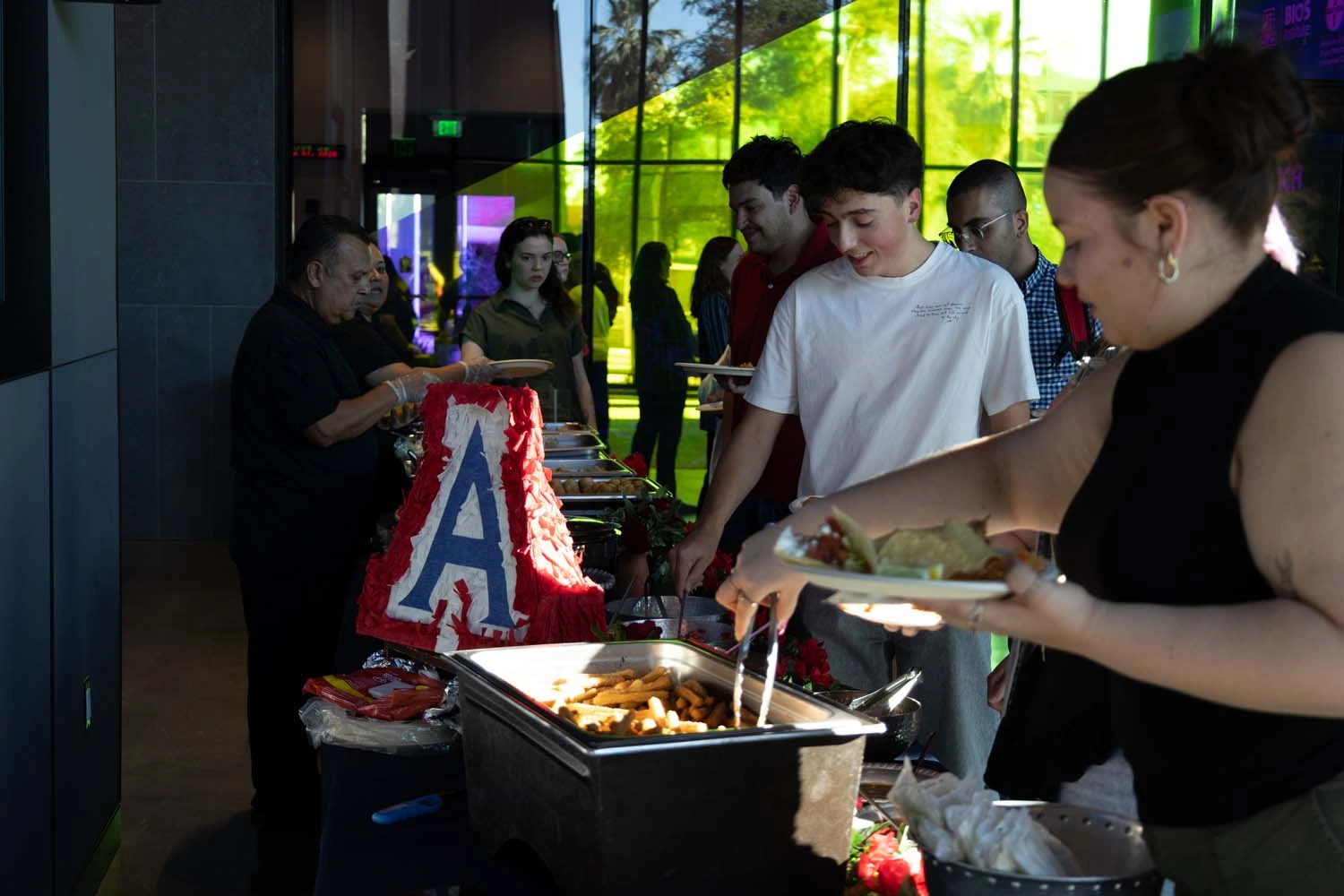 A group of people standing in a buffet line, serving themselves food from trays. There is a cake with a large letter "A" displayed prominently.