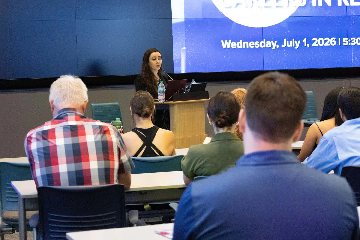 A person stands at a podium giving a presentation in a conference room, with an audience seated at tables. 