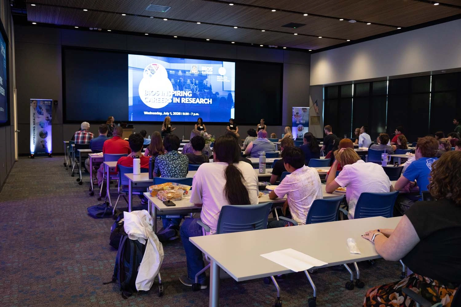 A conference room filled with attendees seated at tables, facing a panel on stage. 