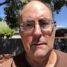 Close-up of an individual wearing glasses, outdoors with trees and a sunny sky in the background.