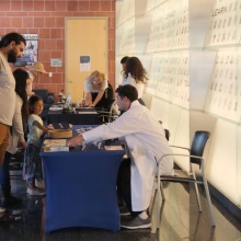 A person interacts with visitors at the Bios Institute booth inside a building with a wall featuring numerous etched portraits and inspirational words.