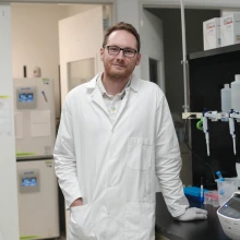 A person in a lab coat stands in a laboratory, leaning against a counter with pipettes and research equipment visible. 