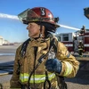 A firefighter in full gear stands in front of a fire truck labeled "Fresno Fire," with industrial buildings in the background. The firefighter is holding a helmet under the left arm and a radio in the gloved right hand.