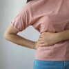 Person in a pink shirt holding their lower back, possibly indicating back pain, against a plain grey background.