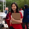 Young person in a red shirt smiling and holding a clip board with an event occuring in the background
