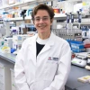 A person wearing a lab coat with a "BIO5" and a University of Arizona logo stands smiling in a laboratory filled with scientific equipment.