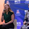 Two individuals seated on stage, speaking at an event at the BIO5 Institute, The University of Arizona. The person on the left holds a microphone and is speaking, while the person on the right listens attentively.