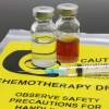 Two vials of chemotherapy drugs, one clear and one amber-colored, beside a syringe on a yellow caution mat labeled "Chemotherapy Drugs: Observe Safety Precautions for Handling."