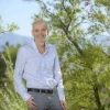 A person standing outdoors, smiling, with blooming trees and a mountainous backdrop. The individual is wearing a patterned blue shirt and gray pants.