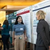 Two individuals discussing research posters at an academic conference, one smiling and engaging with the other who is attentively listening. The backdrop features detailed scientific posters with charts and text.