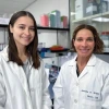 Two researchers in lab coats smiling in a laboratory, with shelves stocked with lab supplies in the background. 