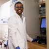 Person in a lab coat and gloves smiles while standing beside a microscope in a laboratory setting.