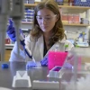 A person in a lab coat and safety goggles is using a pipette in a laboratory setting. The background features shelves with various lab supplies and equipment.